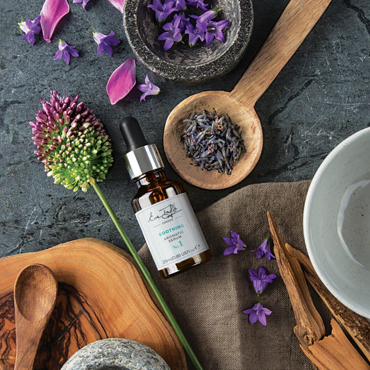 Herbal elements and a bottle of serum on a dark surface with wooden spoons and bowls.