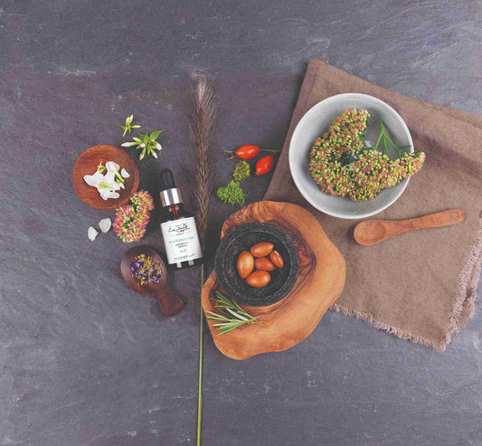 Serum bottle on a dark surface with various natural items and a bowl of flowers.