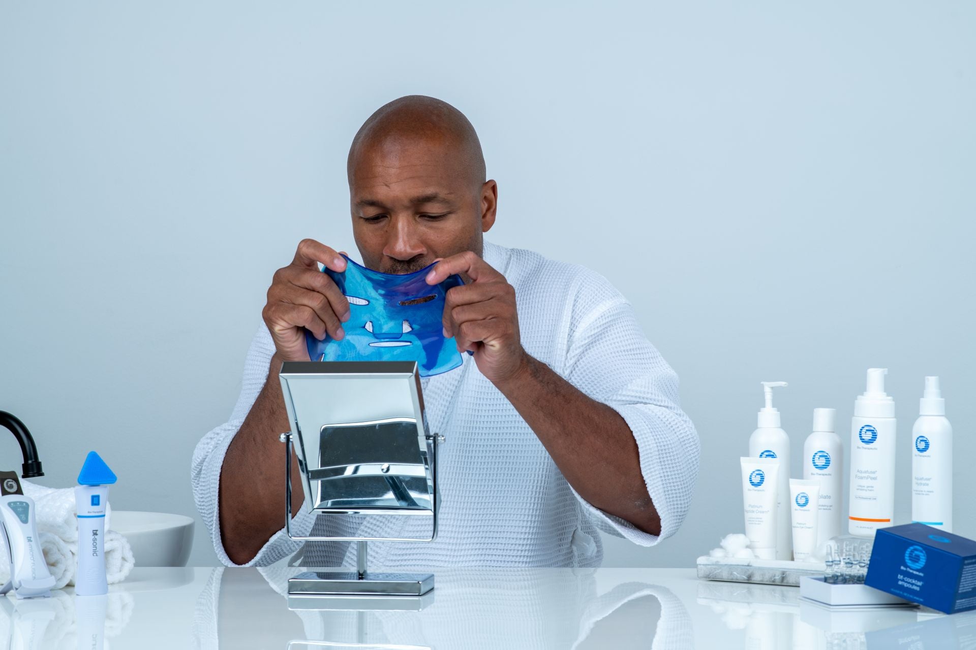 Man holding a blue face masque near skincare bottles and a mask tool tray